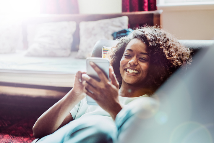 Happy woman using mobile phone while laying on sofa