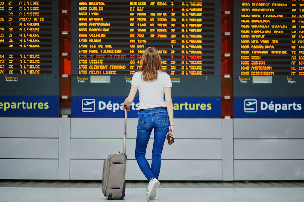 Una mujer joven en un aeropuerto internacional con equipaje y pasaporte mirando la pantalla de información del vuelo