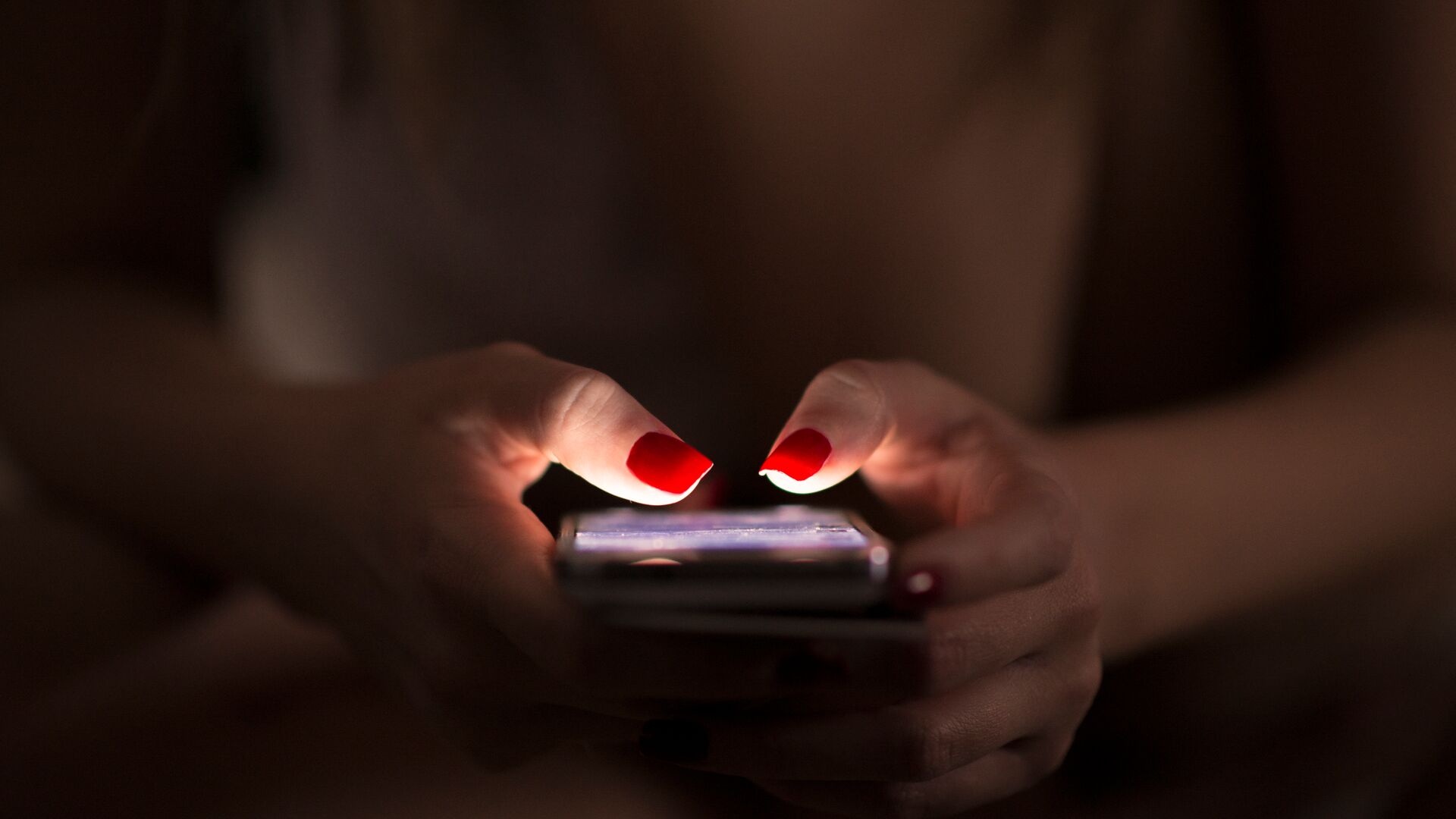 A woman using a cell phone in the dark. Her fingernails are painted bright red.