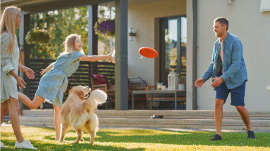  Father, dog & 2 teenage daughters playing Frisbee in their yard.