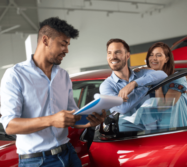 Una pareja feliz negocia el alquiler de un coche nuevo con un vendedor en un concesionario. El coche es rojo.