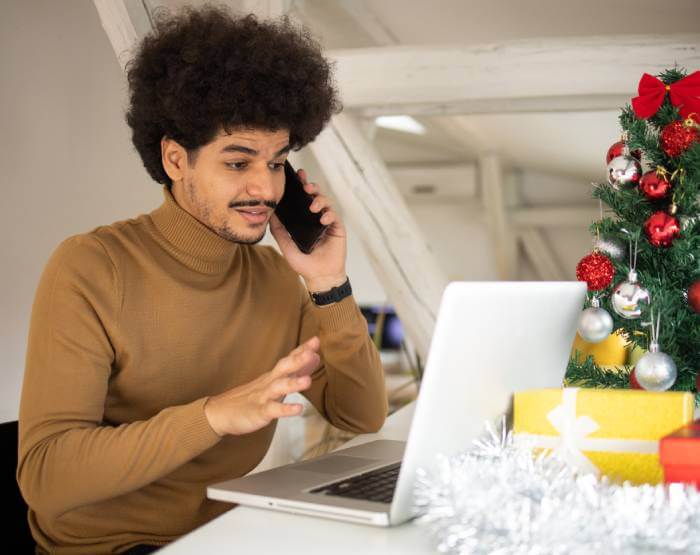 Stressed man talking on phone and looking at his laptop screen hoping he hasn't been scammed. A Christmas tree is in the background.