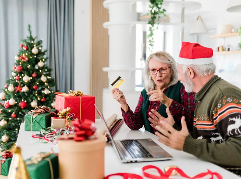 Older man and woman during Christmas season sitting in front of a laptop. The woman is holding a credit card for an online purchase. A decorated Christmas tree is nearby.