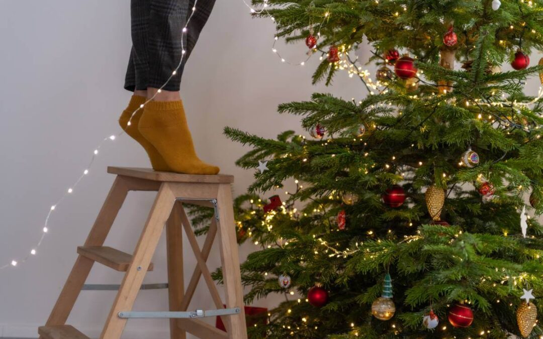 Mujer parada de puntillas en la parte superior de una escalera colgando luces en un árbol de Navidad.